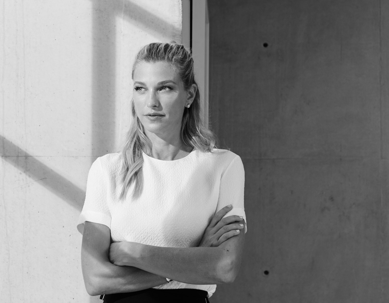 Nicole Junkermann standing with her arms folded beside a sunlit concrete wall, a black-and-white portrait